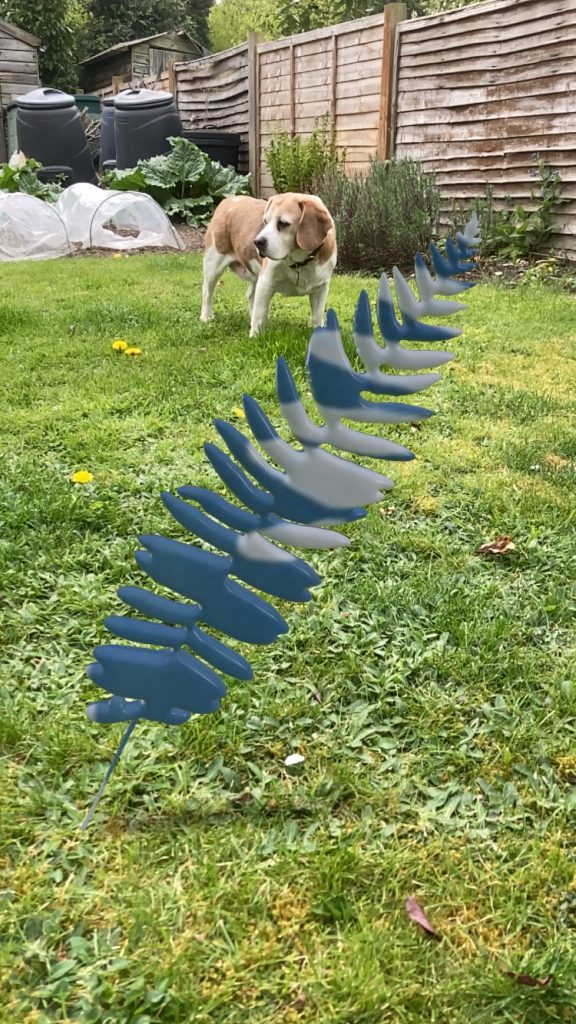 One of Anna Atkins leaves created in Adobe Illustrator and Vectary with the Anna Atkins print used as a material. Photo is of the leaf placed over grass with a fence and a dog in the background.