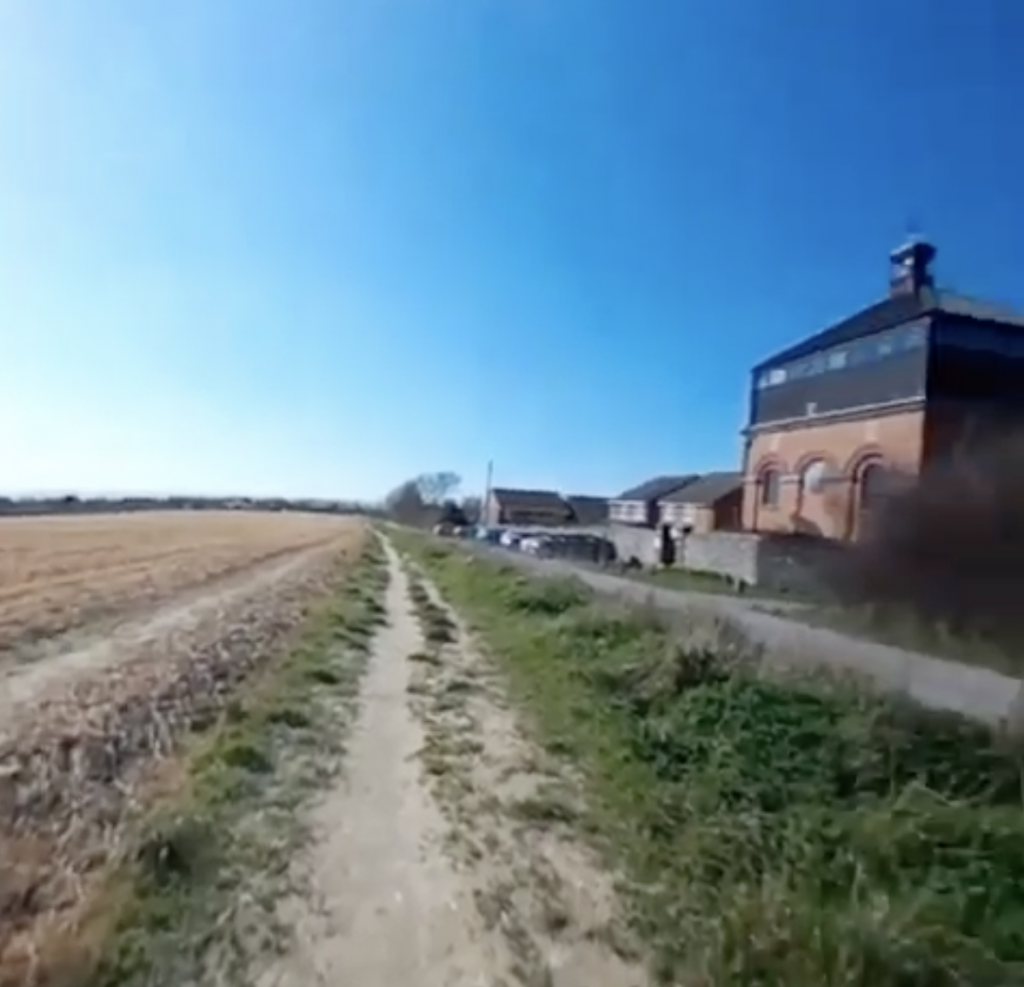 Screenshot of Foredown Tower. There is a dry field to the left of the image, path in the centre, then the road that goes alongside Foredown Tower on the left of the image. Foredown tower is an old water tower and so is a cube shaped made of bricks with a black triangular roof. 