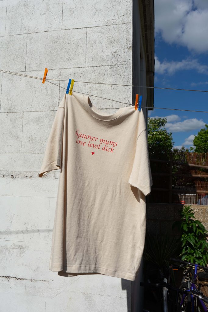 This image is a colour digital photograph in portrait orientation mode. The image focuses on the off-white t-shirt with the graphic design described previously: “hanover mums love level dick” in lower-case chancery cursive font and a little heart below the text – all in red ink. The t-shirt hangs on the laundry rail and is set against the background of a white terraced house wall, a perfect blue sky with chunky clouds, a backyard fence and some bikes stored by the wall.