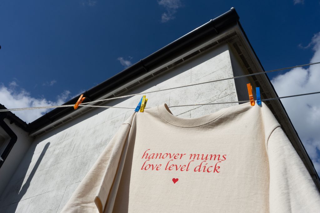 This image is a colour digital photograph in landscape orientation mode, showing the same set-up with the t-shirt hanging from the laundry line. The photograph is shot from a low angle, the camera is looking sharply upwards. At the forefront is the upper part of the t-shirt with the “hanover mums love level dick” text, and in the background is the wall and the roof of a classic Hanover terraced house. The corner angle of the house roof juts towards the sky.