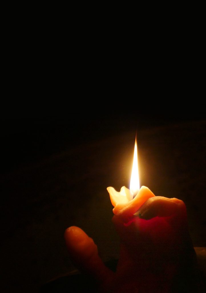 A dark black landscape image is present. In the bottom right hand side of the image is a flame burning from a semi curled wax hand. We can faintly see the tips of some names and some fingers. It is evident the wax is melting. 

This hand belongs to my ancestors.