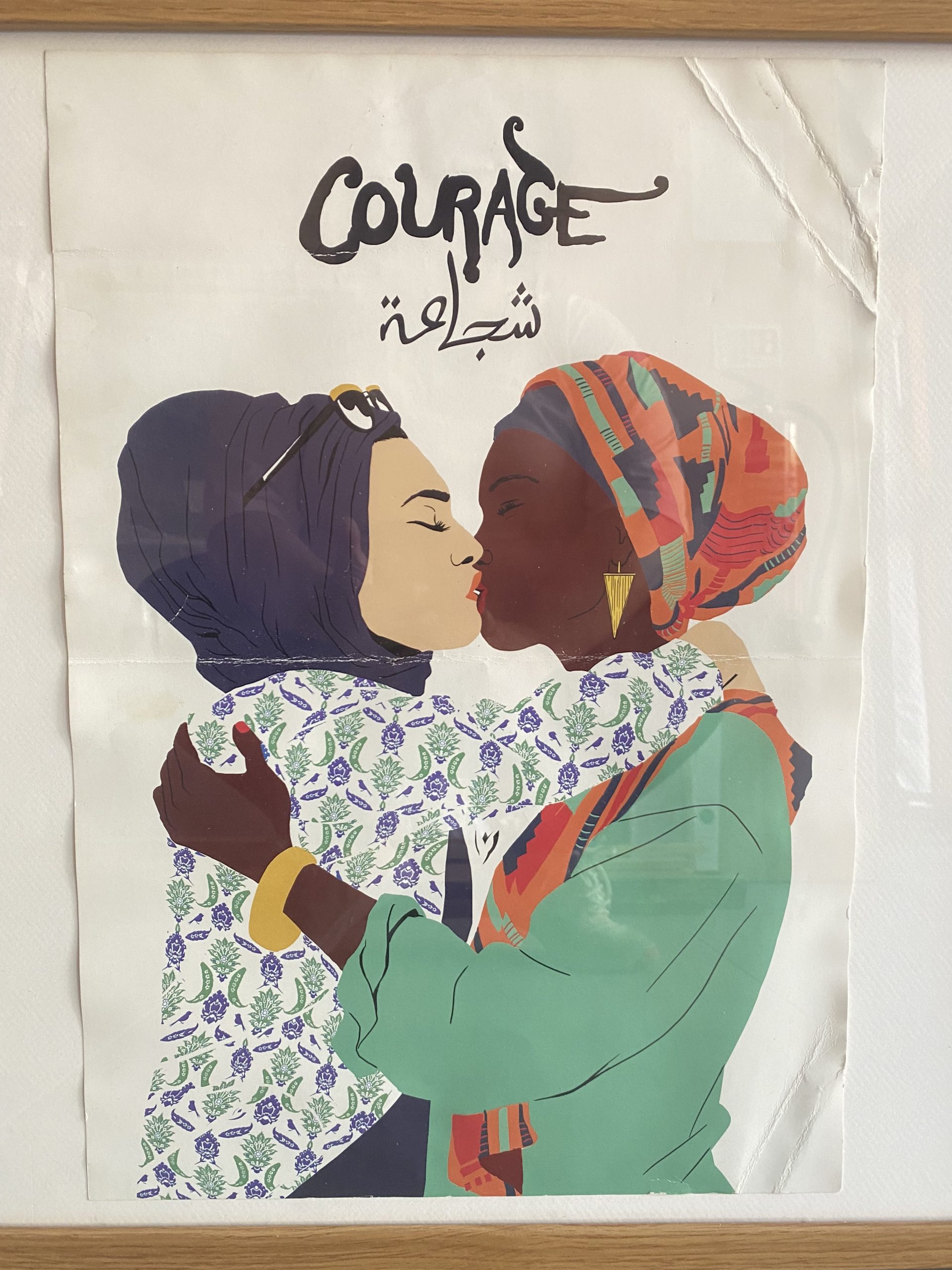 a poster of two women kissing. 
the one on the left is a light skinned woman in a hijab with sunglasses on her head. her eyes are closed and her lips and intertwined with a black woman who is on the right hand side of this poster. the black woman is wearing a fabric around her head also. they are embracing each other and holding each other lovingly. at the top of the poster is one word in two different languages. COURAGE in English and Arabic. In Arabic it is pronounced 'shaja-ah'