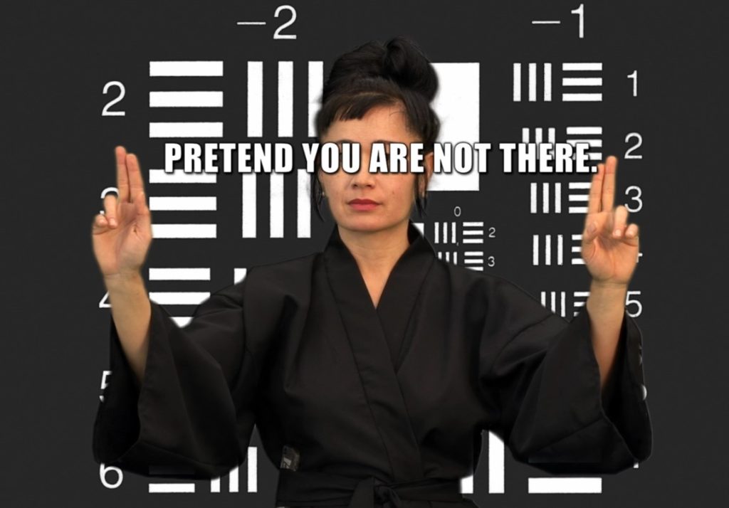 Woman holding up her two fingers on both hands with a text in between her arms reading 'Pretend You Are Not There'. The background is dark grey with white rectangle shapes and numbers.