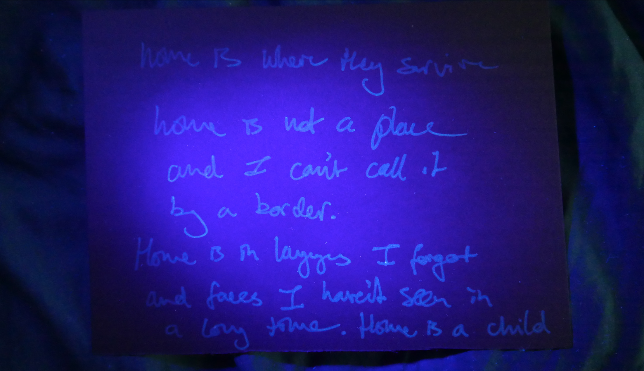 Image of a black piece of card with an anonymous contribution written in invisible ink only seen here through a UV light. The contribution is a hand written note from a member of the general public in London UK, in response to the question 'what does home mean to you?'. Under purple light, the faint text reads "home is where they survive. home is not a place and I can't call it by a border. Home is a language I forgot and faces I haven't heard in a long time. Home is a child"