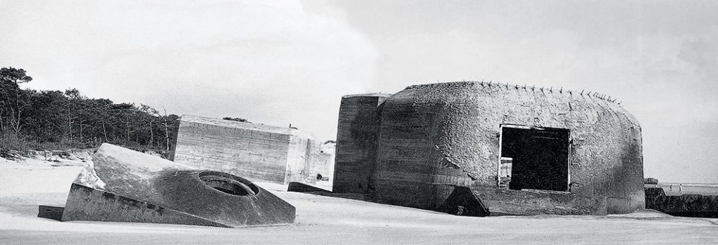 A black and white image of a rounded concrete bunker emerging from a sandy beach. One circular opening leads into the ground.