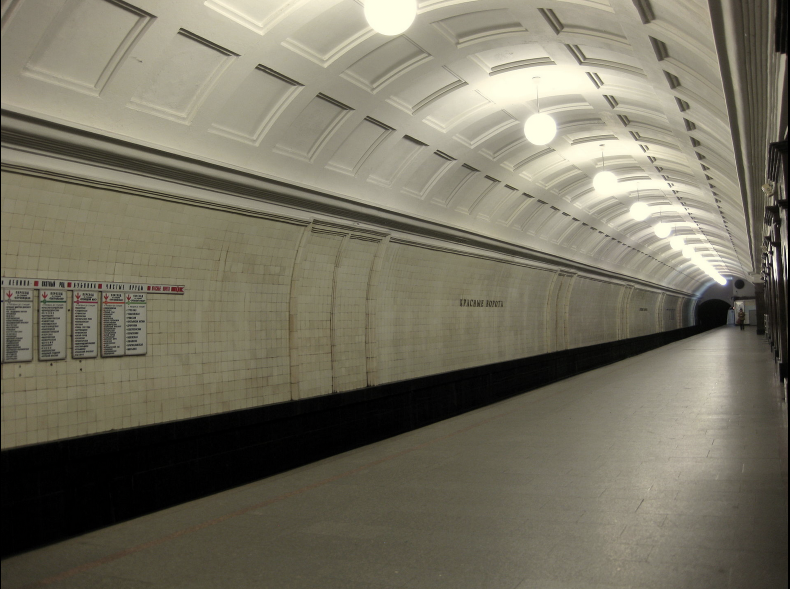 This image is a photograph of Krasnye Vorota Metro Station. It shows a creme-coloured vaulted ceiling with tiled walls. It bears a striking similarity to Gants Hill station in London. In a small difference between the two, this image shows orb-like lights hang from the ceiling. The platform is empty of people and the angle of the photograph emphasizes how barrel-shaped the station design is. 