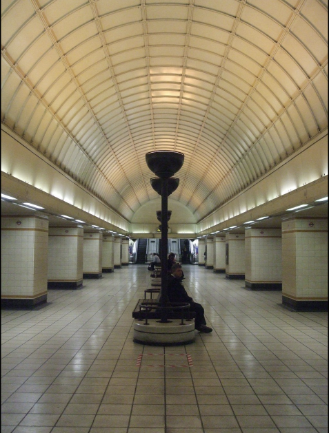 This is an image of Gants Hill train station showing the central concourse. It shows a creme-coloured vaulted ceiling with a row of benches through the middle where some commuters are sitting. At the end of the shot, small but visible in the distance are the escalators going upward. The station is lit by a row of tall upturned lamps across the cental line of benches. The photograph is beautifully symmetrical, except for the commuters who predominantly face the platform leading into central London.