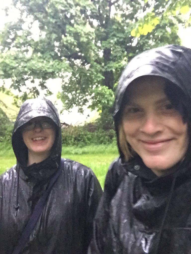 A photograph of me and my girlfriend, Maria, with adorable matching black raincoats, smiling like a pair of fools, utterly drenched by the rain. The green of a field and some trees can be seen in the background.