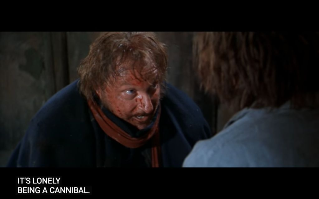 A medium-shot image of an unkempt white man with ginger hair and beard, looking very muddy and spotted with blood, with bulging eyes look up towards a character slightly out of shot, over whose shoulder we see the ginger haired man. Below this are subtitles that read: "it's lonely being a cannibal"
