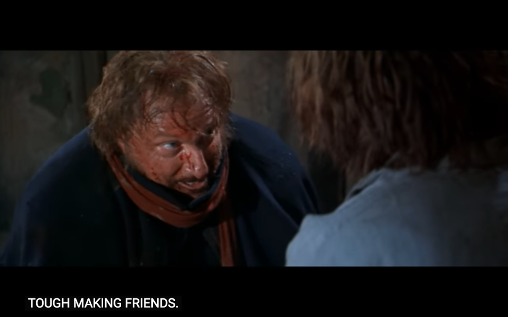 A medium-shot image of an unkempt white man with ginger hair and beard, looking very muddy and spotted with blood, with bulging eyes look up towards a character slightly out of shot, over whose shoulder we see the ginger haired man. Below this are subtitles that read: "tough making friends"