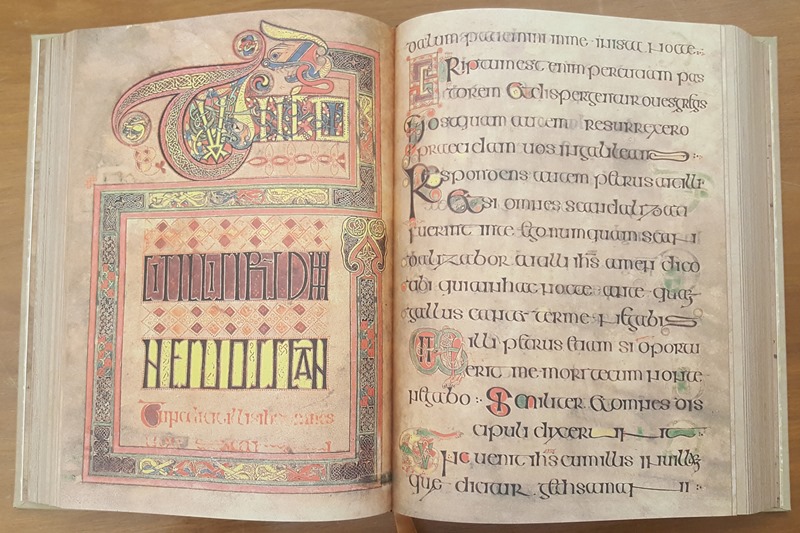 An image of a book spread open. The book is the Book of Kells. On the left hand page is a square of celtic knots words in Latin written in a font that makes it hard to read. Above this square is word in an ornate script. The colour pallet is red, blue and yellow, and the paper looks very aged. On the right hand page is a page of writing in Latin, with the first letter of the first word of each sentence being drawn in a coloured flourish, whilst the rest of the letters in all the other words are black. 