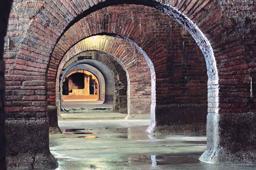 A series of brick semi-circular tunnels disappear into the distance, where stairs can be seen ascending out of these depths. The smooth floor is covered in puddles of water. This is an Ancient Roman sewage system that is light enough that it appears it is now used more for exhibition and tourists than for everyday use.