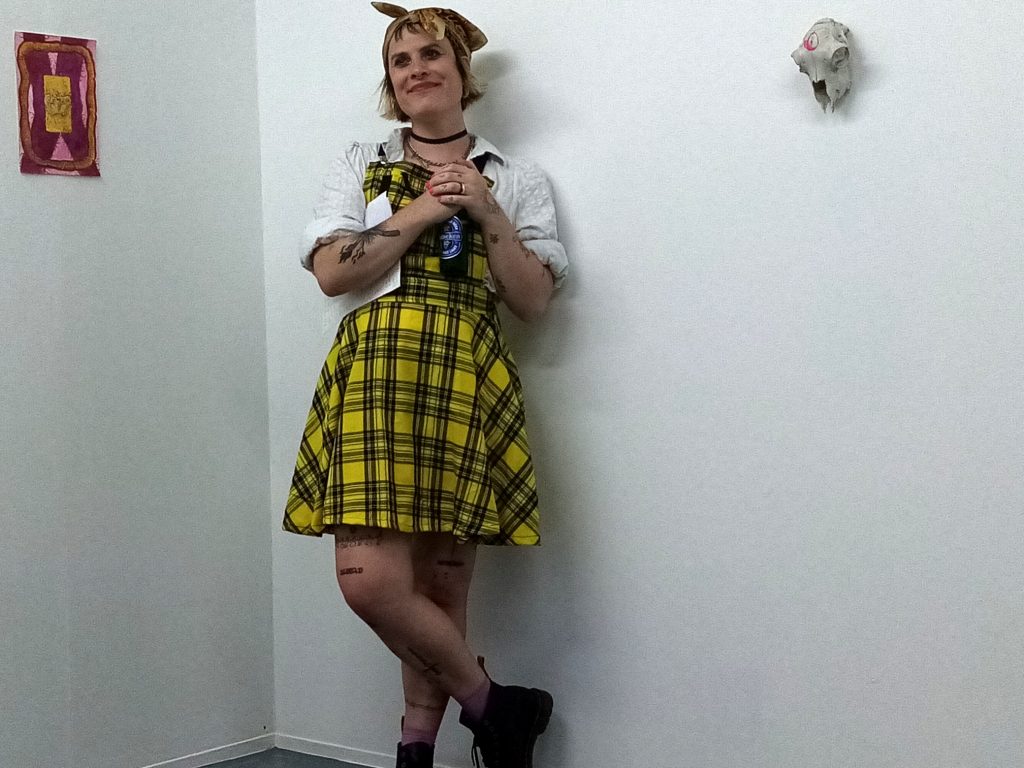 Photo of Sammy Paloma wearing a yellow and black tartan dress and white blouse standing in the white corner of a gallery space.