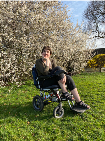 A digital photograph (portrait, colour) of what looks like someone sat in a powered wheelchair on a sunny day. They are turned to the camera smiling softly with one knee lifted and their head on their hand. They are in a grassy field with a large bush of spring flowers behind them. The person has pale, white skin and rosy, round cheeks with short brown hair. They are wearing black trousers, black sandals and a green long sleeve top. The image is nice and the setting sweet.