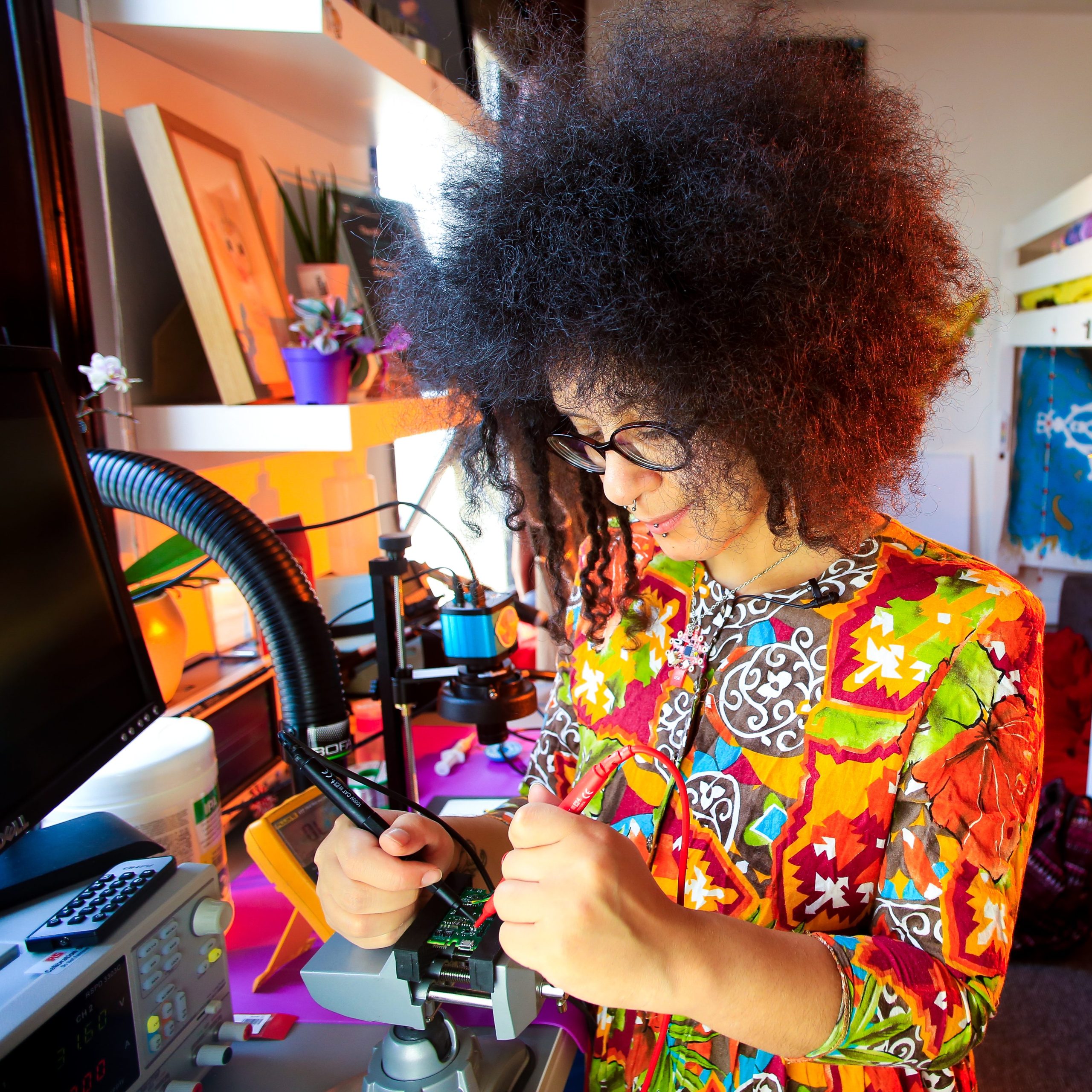The image shows Shrouk working intently at their colourful and creative electronics lab. Shrouk has an afro hairstyle and is wearing a brightly coloured dress which is mainly deep oranges and reds with intricate patterns. She is working on a printed circuit board with an oscilloscope, seemingly deeply focused on the detailed electronics work or experiment they are engaged in.

Shrouk's workspace has an artistic, energetic vibe, filled with various bright decorations and objects in oranges, pinks, blues and greens that match Shrouk's expressive and unique style. The photograph captures a moment of concentration as Shrouk applies their skills to an electronics project, with the vibrant atmosphere of the lab reflecting Shrouk's creative personality and approach. The setting suggests Shrouk is bringing an imaginative flair to their electronics work.