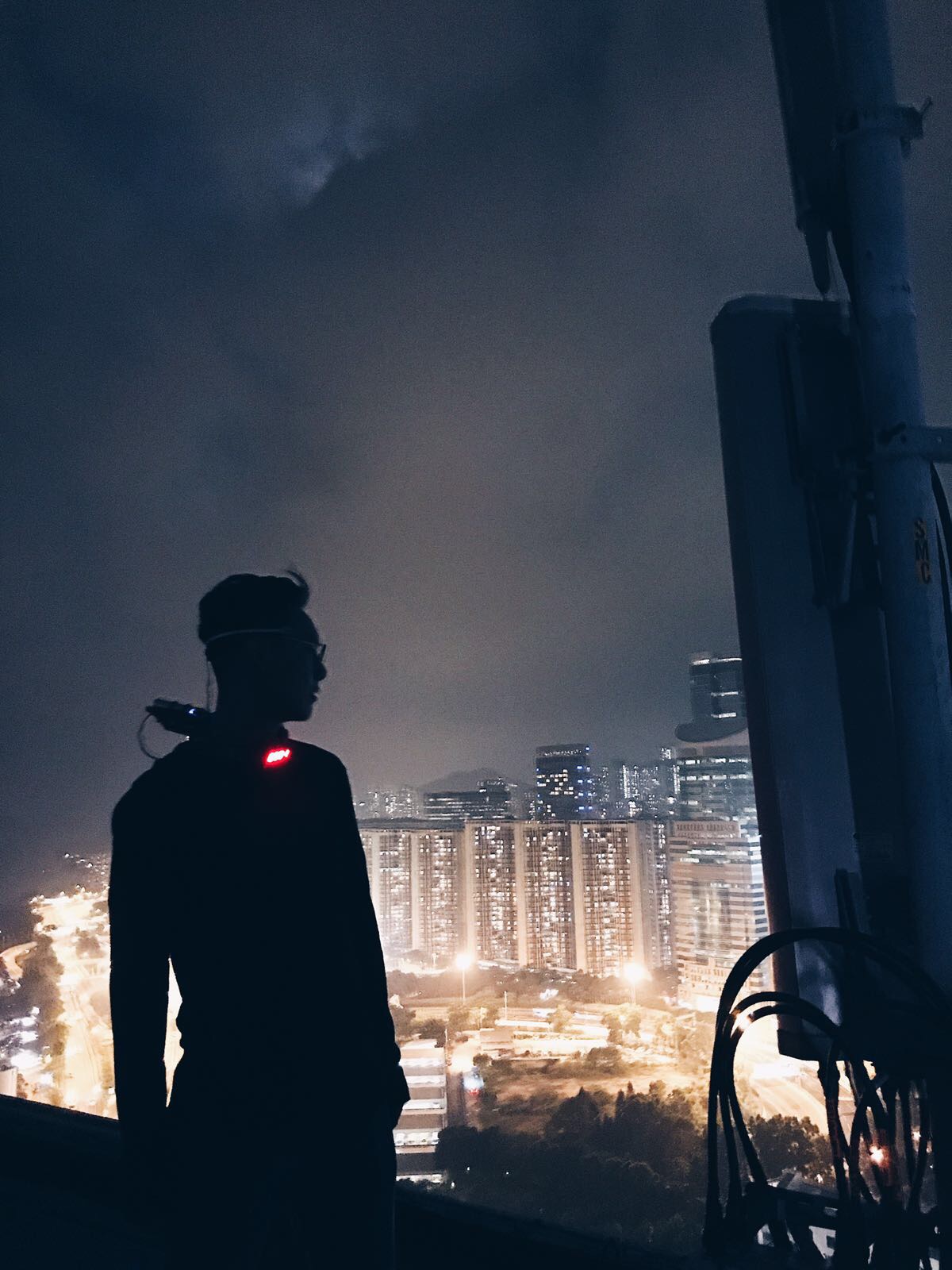 The image depicts a silhouette of a person standing on a high vantage point overlooking a city at night. The person appears to be wearing a headlamp or some device with a red light on their back. The cityscape below is illuminated with streetlights and lights from numerous tall buildings. The sky is overcast with clouds, adding to the dramatic atmosphere of the scene. In the foreground, there is a large metal structure, possibly part of a communications tower or similar equipment.