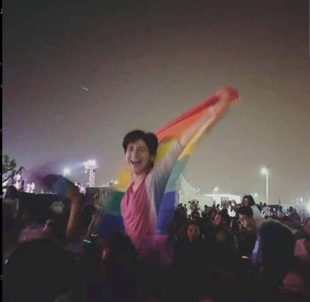 A photo of Sarah Hegazi smiling widely and holding a rainbow flag aloft at a crowded nighttime outdoor event, with stadium lights visible in the background.