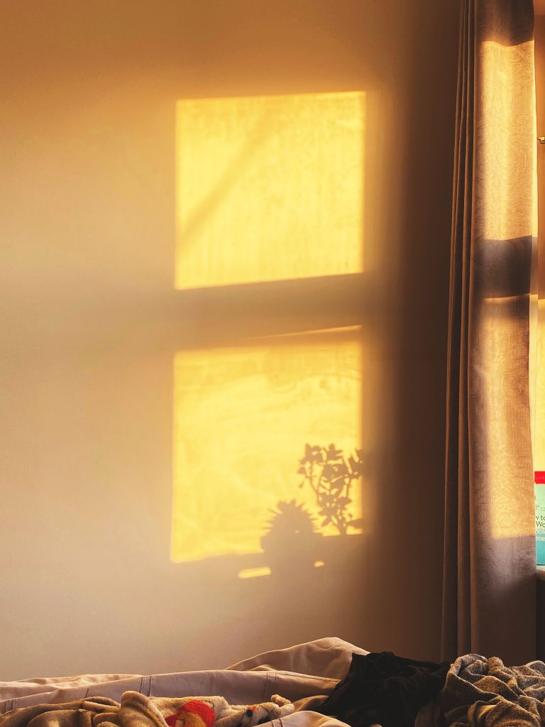 Warm golden sunlight streams through a window, casting the shadow of the window frame and two potted plants onto a beige wall. The shadows are clear and rectangular, creating a peaceful, minimalist composition. Part of a bed with soft, rumpled blankets and clothes is visible at the bottom of the image, adding a lived-in, cozy atmosphere. A curtain hangs partially open to the right, revealing a sliver of the window.