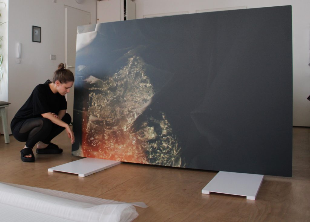 Leah crouching down next to a very large dibond photograph. She's a white woman with dark blonde hair up in a bun, wearing all black. The photograph is 2metres tall and 3metres wide, and rests on white foam blocks. It is mostly darkness and shade, but at its left side glows into a warm red flare, over rippling sunlit water. The room might be a studio or domestic dating, with wooden floors and a closed door, framed photo on the wall, and a door buzzer phone on the left.