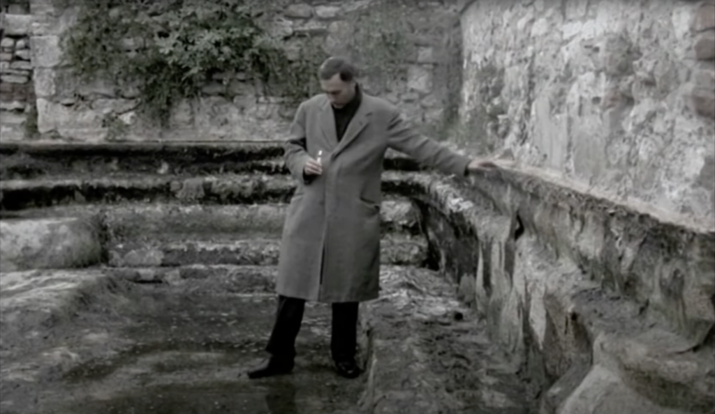 The same man in the same coat, but dry. This image is more desaturated, the colours are dull. He stands at the bottom of an empty and somewhat ruinous stone pool. He is touching its side, holding a lit candle, looking down at it and focusing on its little flame as he starts to take a step away from the wall.