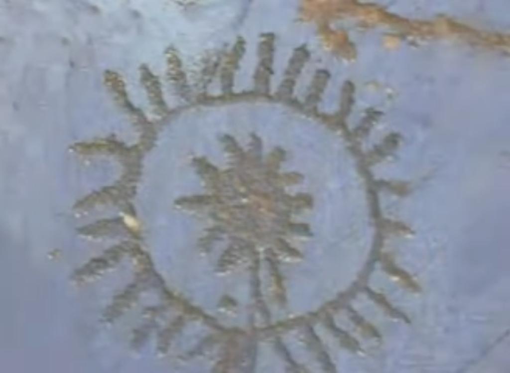 A low-res image of a roughly drawn, or maybe carved, golden sun shape with protruding rays, and a corona around it with more rays. The surface it’s carved into appears to be white or pale blue stone.