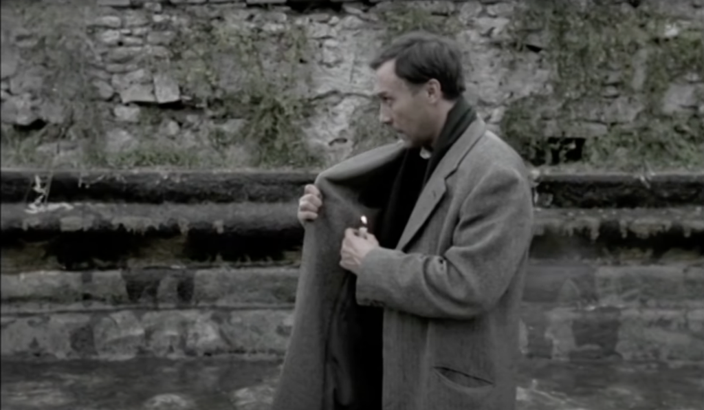 He has given up on cupping his hands around the flame, and is trying to shelter it from any breeze with the inside of his coat, which he holds next to it.