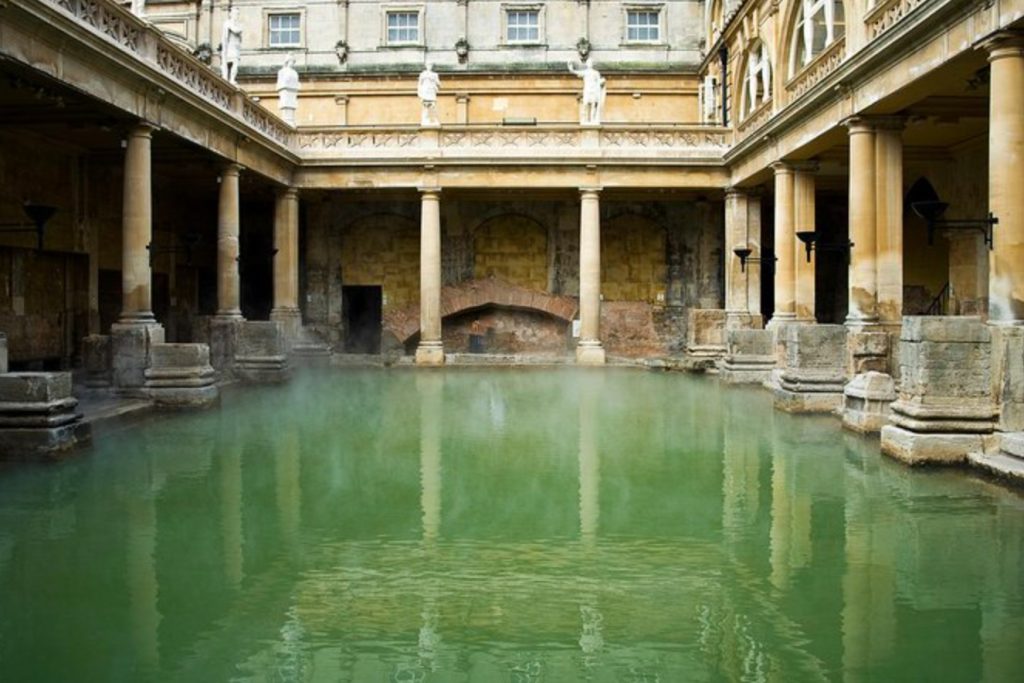 Daytime, a central view across a green rectangular body of water, Roman pillars surrounding it. A small walkway runs behind the pillars, ending at ancient walls. Above, statues line a balcony, and the windows of other old (but perhaps less ancient) buildings surround it.