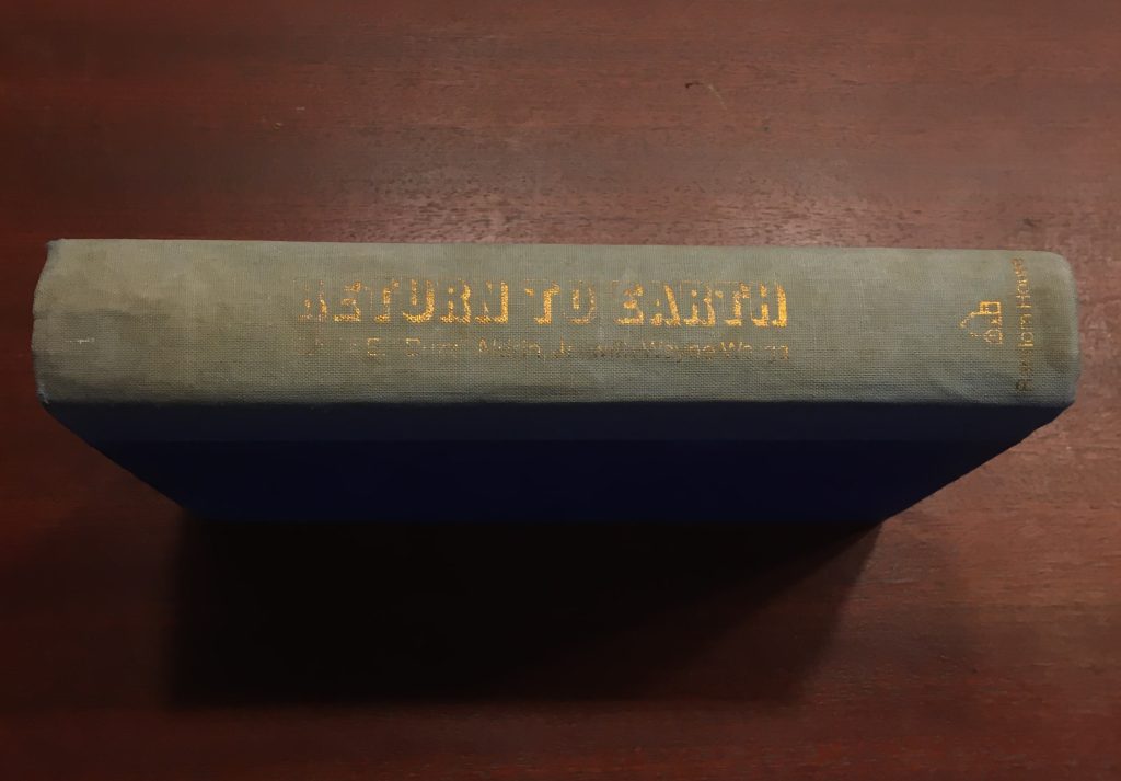 A book standing spine-up on a dark cherry wood surface. It is old and beaten up, worn-out blue, with faded gold lettering that just about reads: RETURN TO EARTH. More text underneath and to the side are illegible. It casts a monolithic shadow in front of itself.
