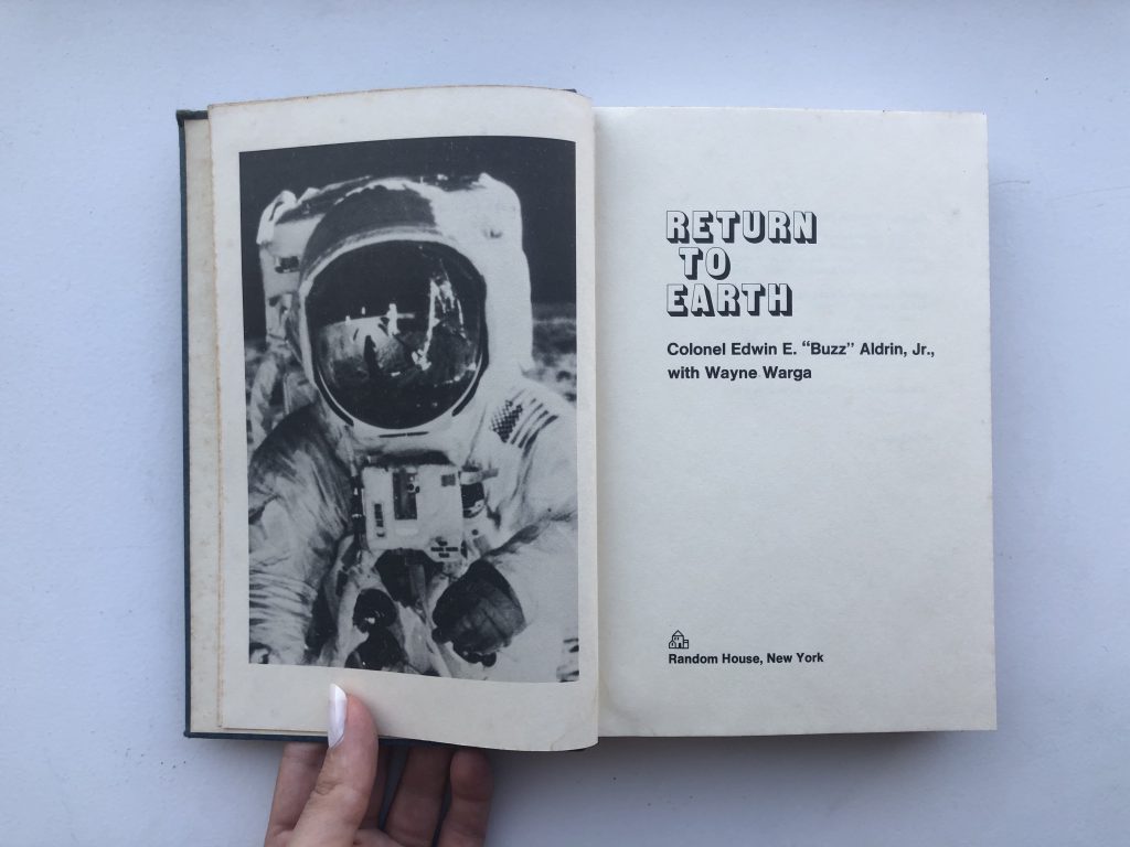 An open book. On the left page is a black and white photo of an astronaut in their suit. In the reflection of their helmet is the sketchy figure of someone else, or perhaps just some equipment. The horizon in the reflection and behind the astronaut is black, meeting the edge of the moon they’re standing on. On the right page is text in a dated font which reads ‘RETURN TO EARTH, Colonel Edwin E. “Buzz” Aldrin, Jr., with Wayne Warga, Random House, New York.’ A white hand holds the book open, a milky painted thumbnail pins down the left page at the bottom. The book is resting on a white surface.