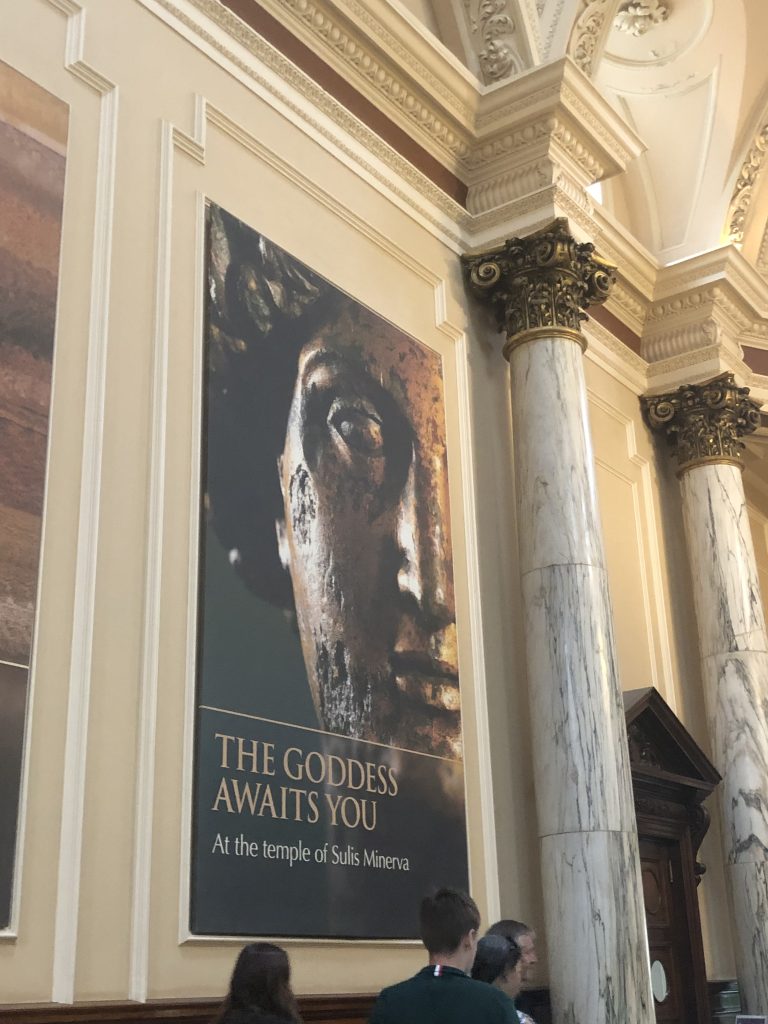 A museum banner in a pillared 18th century interior. On it is a Roman sculpted face, obscured on shadow and picked out on dramatic light. It reads ‘the goddess awaits you at the temple of Sulis Minerva’