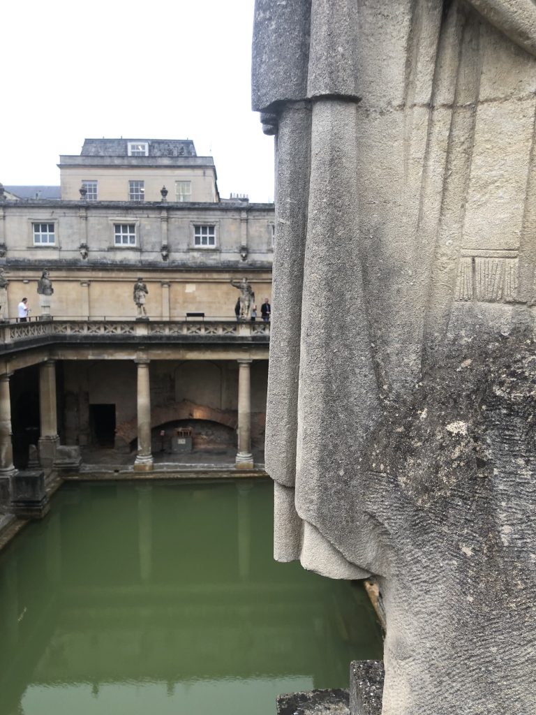 Behind a statue, its cape hanging in folds, we look down upon the green bath from a height. The statue’s counterparts face it opposite, along the walkway which follows around the edge of the bath. Each of them is permanently posed, guarding or adorning the watery centre.