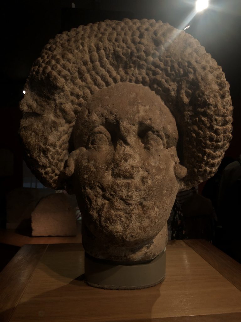 A stone sculpted head in a dark space. It’s on a plinth and its mouth and nose have eroded away, leaving blank eyes and and impressive plaited hair arrangement like a crown over her head.