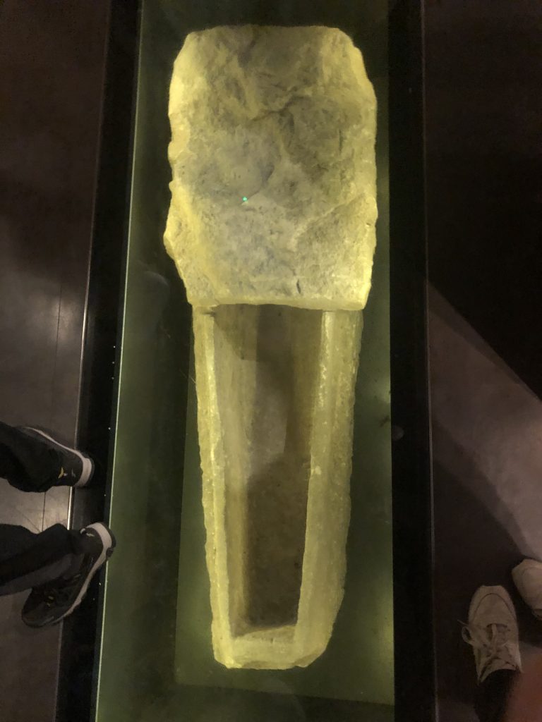 A coffin underfoot, under glass. It is small and yellowish, made of an unknown material. It is enclosed at one end, and warped by its two thousand years.