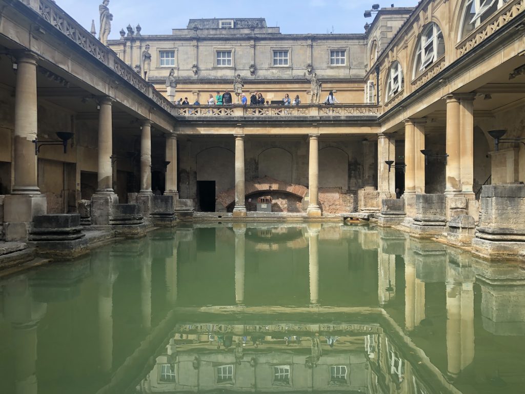 A central view of the bath from the bathside: a green rectangular body of water, Roman pillars surrounding it. A small walkway runs behind the pillars, ending at ancient walls. Above, statues line a balcony, and the windows of other old (but perhaps less ancient) buildings surround it.