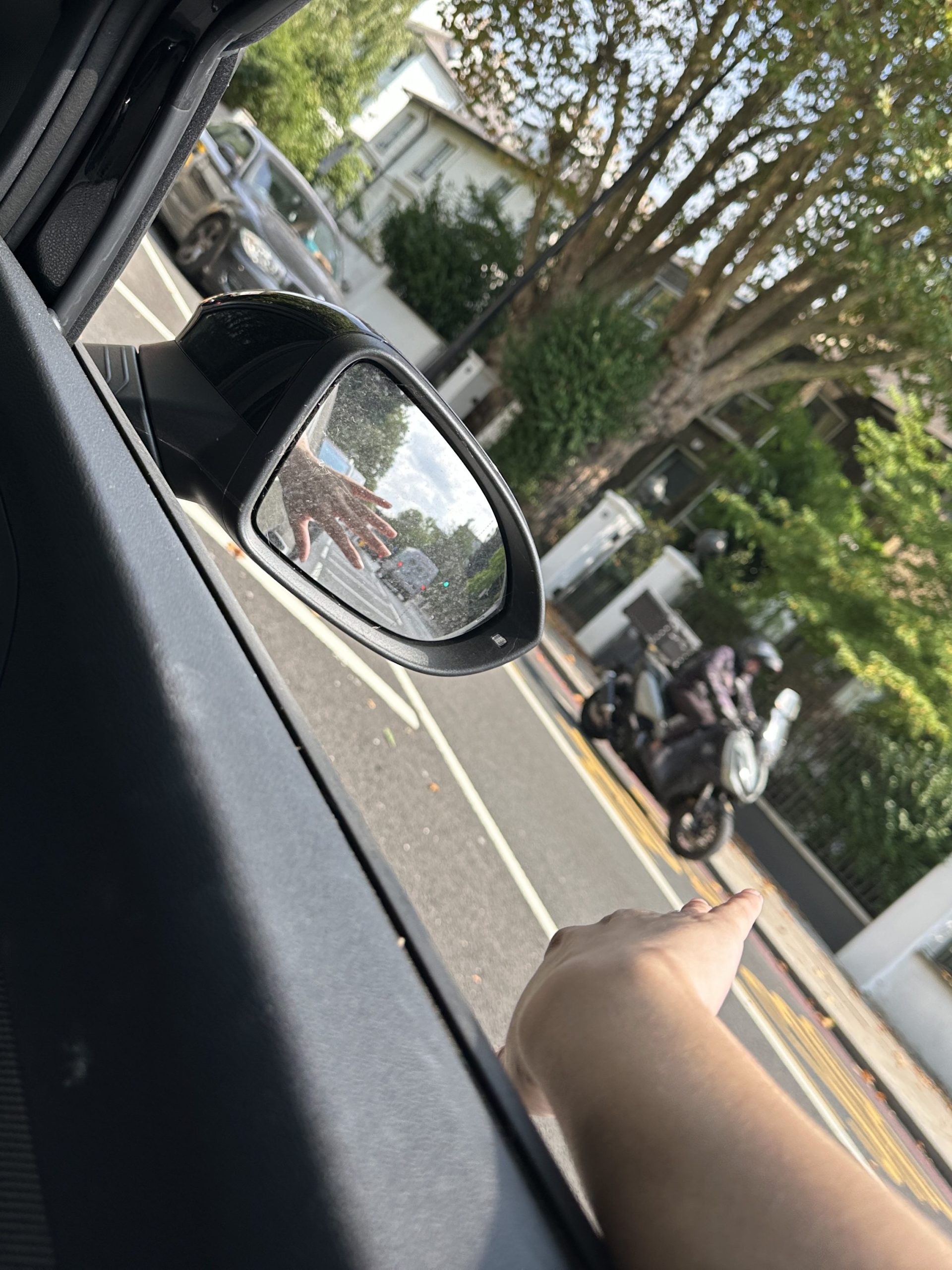 An arm hangs out a car window t on a tree-lined street In London. The car's side mirror reflects the hand feeling a gentle breeze. It is a sunny day.