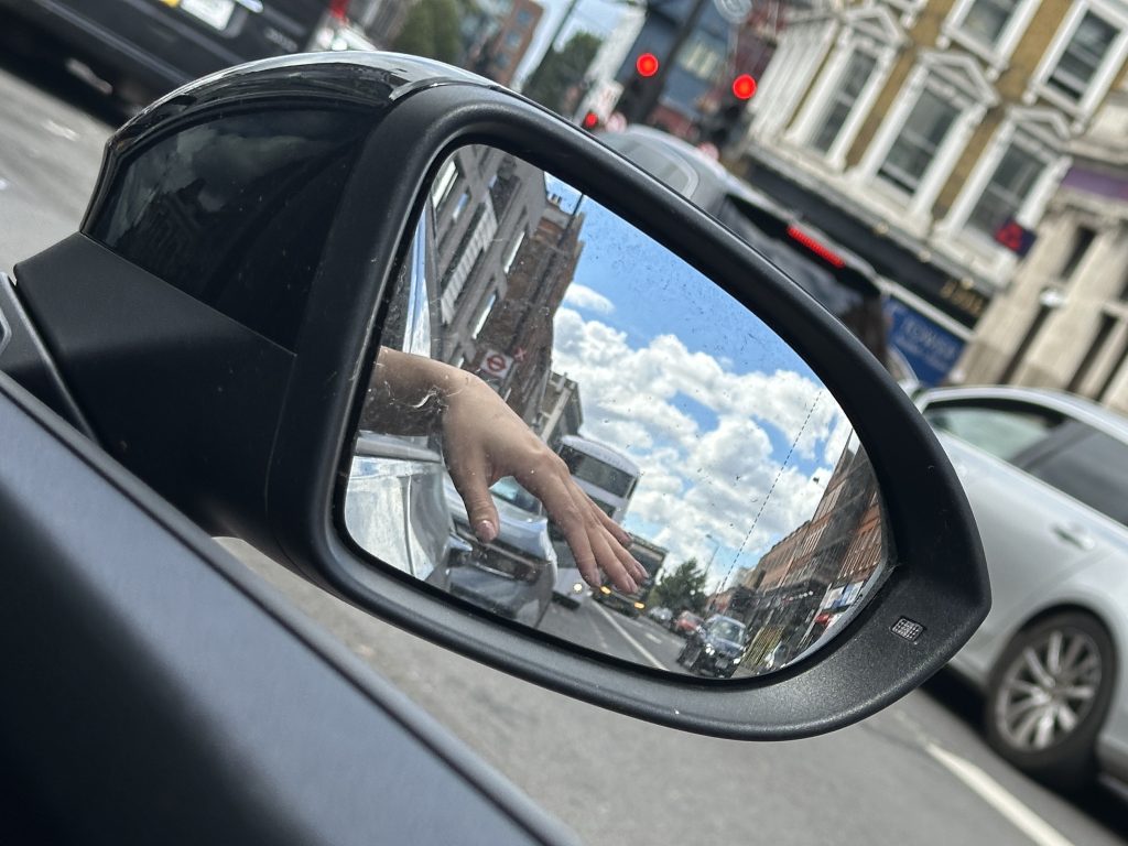 A hand hangs out of the car window and is reflected in the wing mirror. The road behind doesn't look too busy.