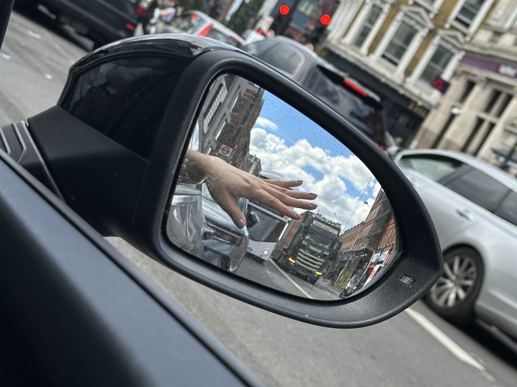 A hand hangs out of the car window and is reflected in the wing mirror. The fingers are stretched out as if trying to reach or perhaps gently asking for attention. The road behind gets busier. There is a double decker bus and lorry approaching.