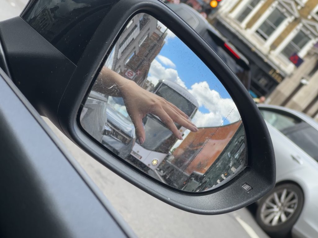 A hand hangs out of the car window and is reflected in the wing mirror. The fingers are straight but relaxed as if trying to gently asking for attention or maybe feeling a breeze. The road behind gets busier. There is a double decker bus and the lorry passes by.