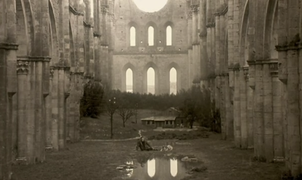 A sepia image. The man is quite distant, a stance somewhere between laying and sitting, arm propping him up. A dog lays next to him, and in front of them is a large puddle. Behind them a small shack and a circle of trees, and surrounding it all, the large ruins of a church.