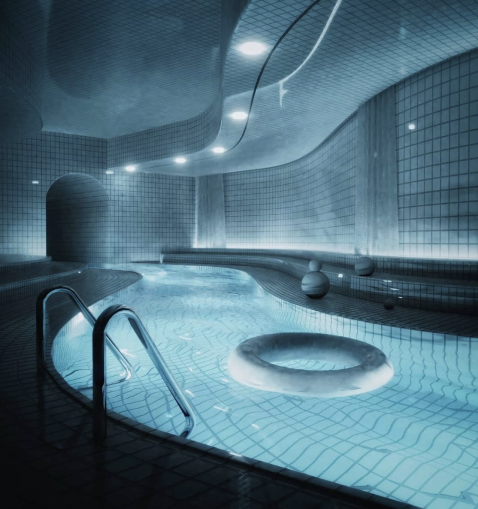 A dark white tiled space with a winding thin pool lit by spotlights from above. A rubber ring floats on its surface next to a metal ladder leading into it.