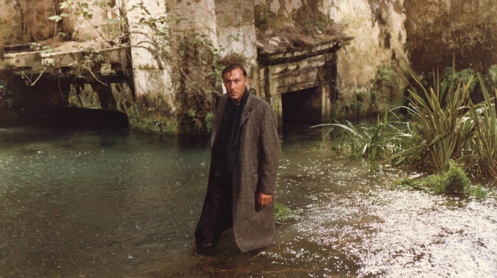 A still from a film shot on film. A white man with dark hair and a long coat stands in a flooded ruin. It’s overrun with plants and algae, and it’s dripping everywhere. The man stands with one hand in his pocket and the other loosely to his side. He looks up and to the left of us intently, as if listening to someone very seriously. The greens are rich against the stone.