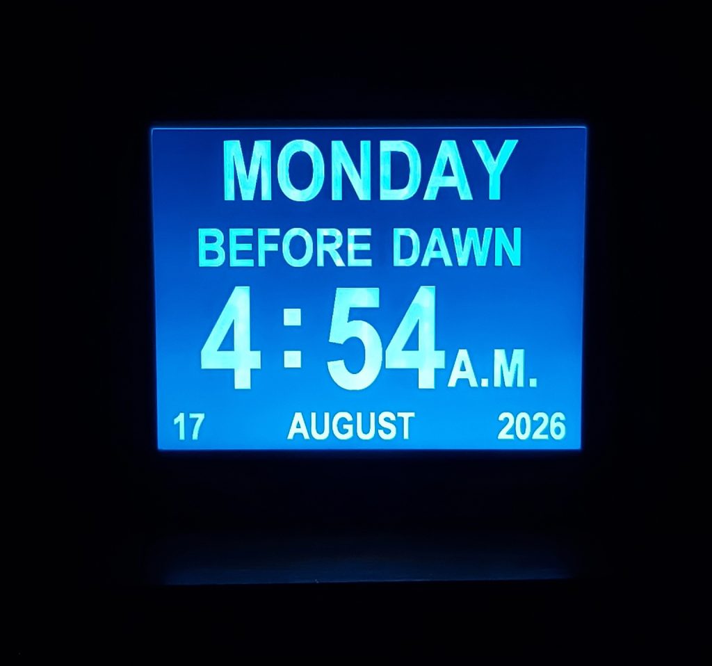 A bright blue digital display set against a black background reads ‘Monday Before Dawn’ in capital letters above the time 4:54 a.m. in larger numbers in the centre of the screen. Running along the bottom of the screen the date reads: 17 August 2026. There is a reflected light from a mantlepiece and the illuminated screen emphasizes the stillness and darkness of the pre-dawn hour.