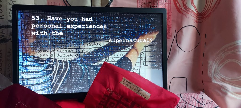 The photograph shows an image of a computer monitor displaying a close-up view of a hand drawing on a hand-drawn chart or grid filled with handwritten text and heavy black lines written in deep black charcoal, making much of the content obscured. On the screen in front of the image white text in typewriter font reads: 53. Have you had personal experiences with the supernatural? behind the screen there is pink and cream fabric printed with hand-drawn circles, lines, grids and dashes in red, white and black lines. In front of the screen, partially obscuring it are machine embroidery charts and diagrams in red stitching on red fabric. 