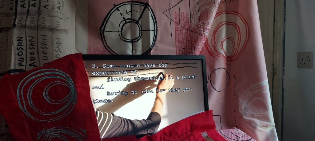 The photograph shows an image of a monitor with a black frame displaying a close-up view of a hand drawing heavy black lines in deep black charcoal, in the image golden early morning sunlight casts a long shadow of the hand across the paper. On the screen in front of the image white text in typewriter font reads: 3. Some people have the experience of finding themselves in a space and having no idea how they got there. Behind the screen there is pink and cream fabric printed with hand-drawn circles, lines, grids and dashes in red, white and black lines. In front of the screen, partially obscuring it are machine embroidery charts and diagrams in red and pale blue stitching on red fabric. Further behind the fabric and the screen on the right there is a view of a doorframe and a plug socket indicating this is a domestic space.