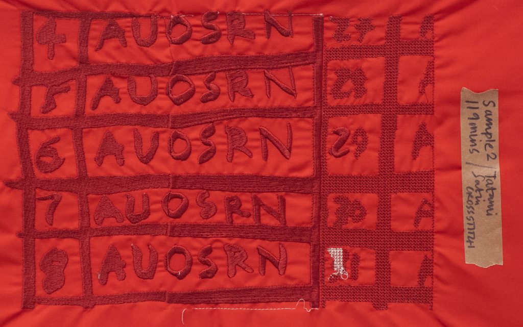 A photograph of deep dark red digital embroidery stitched onto a paler blood red fabric. The embroidery is in the form of a grid with numbers that run from top to bottom 4-8 and 27-31 consequentially and letters that read AUOSRN. The chart is cut off and in places the letters A are cut in half and in others red stitching is over sewn in white. On the right hand side a piece of brown tape with ripped edges has been stuck on the fabric. on the tape hand written text in black felt tip pen reads: Sample 2 119 mins / Tatami Satin CROSSSTITCH.