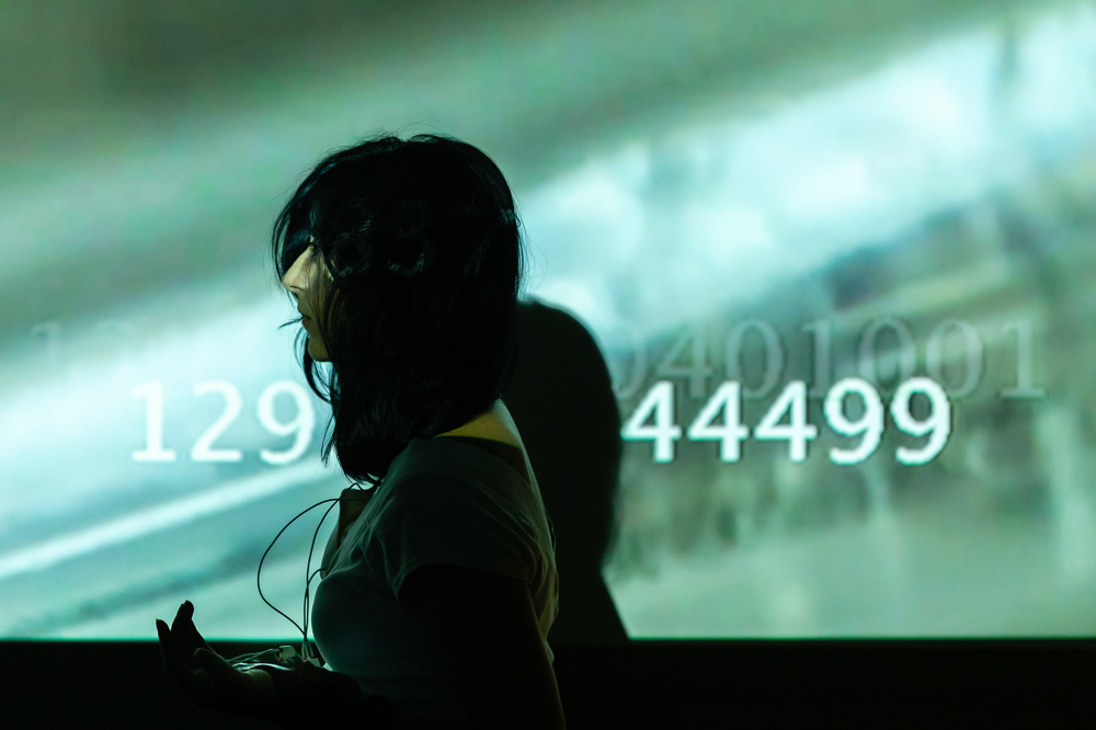 A performer with long dark hair stands in profile before a green-tinted projection displaying blurred imagery and large white numbers. Their face is partly obscured, and their shadow falls across the screen.