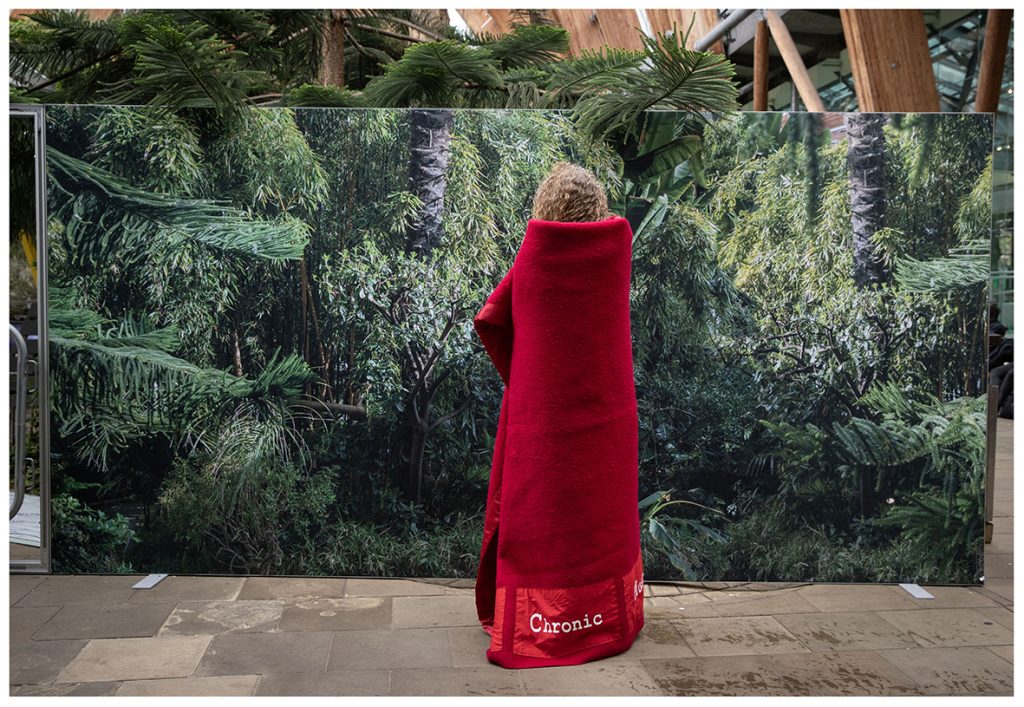 The photograph shows me standing vertically wrapped in a large red blanket. I am facing away from the camera. White typewriter text at the bottom of the blanket reads Chronic. I am standing on a concrete floor in front of a large billboard printed with tropical ferns. Behind the billboard is a large indoor greenhouse with pine trees and wooden architectural structures. 