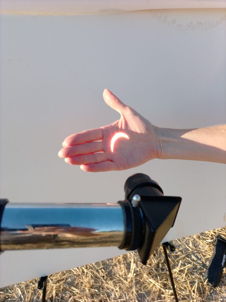 A close-up view of a hand held in front of a white projection screen outdoors. Bright sunlight passing through a telescope projects a glowing crescent-shaped image of a partial solar eclipse in the centre of the palm. In the foreground, part of a telescope is visible, aimed at the screen. The lower edge of the image reveals straw-covered ground, suggesting the scene is taking place in a harvested field. Warm evening light illuminates the hand and screen, highlighting the projected crescent shape, creating a fleeting interaction between body, light, and astronomical observation.