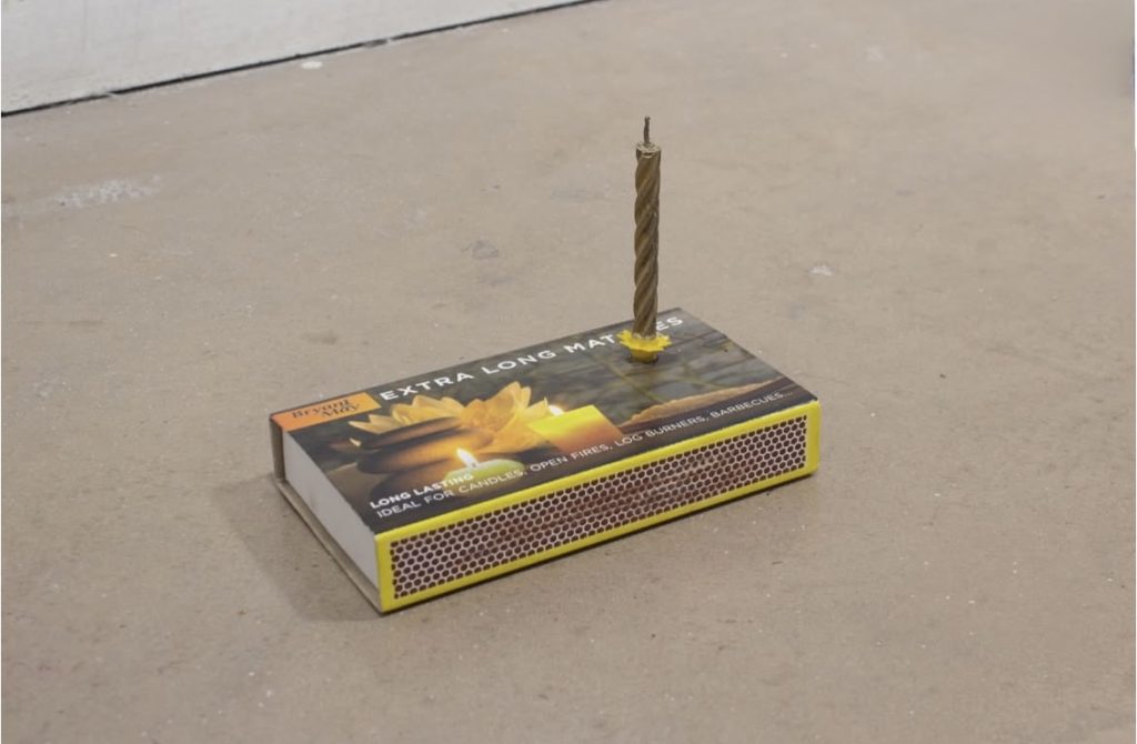A single unlit birthday candle sticks out from the top of a closed match box as if it were a cake.