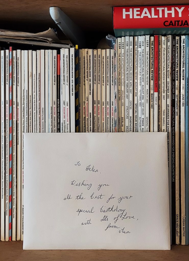 A photograph of an unopened white envelope on a wooden plywood bookshelf filled with architectural magazines and journals. Only the spines of the journals are visible. The envelope sits in the space remaining on the shelf in front of the journals. On the envelope hand-written text reads "To Helen. Wishing you all the best for your special birthday with Lots of Love from Mum." The handwriting is old-fashioned joined up writing with flourishes, written carefully in black pen in the middle of the envelope. In the top right of the image, a thick hardback book rests horizontally on top of the journals, satisfyingly filling the remaining gap between the next shelf. The book is red with the words healthy on the spine in white capitals and Caitja underneath in black capitals.
